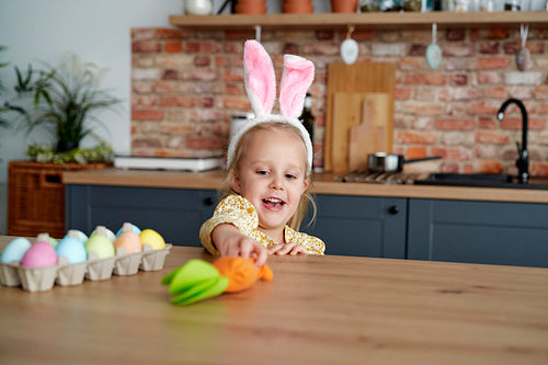 Playful girl in rabbit ears taking a carrot from the table