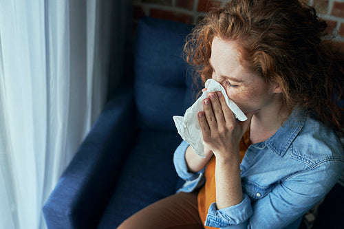 Young caucasian  red head woman blowing nose into tissue at home