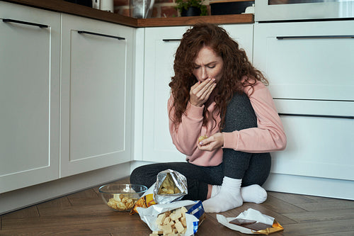 Young caucasian woman eating snacks greedily on the kitchen floor