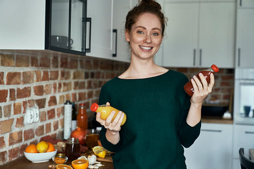 Portrait of cheerful, young caucasian woman in the kitchen, holding two bottles of fruity juice