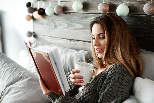 Woman drinking coffee and reading book on bed