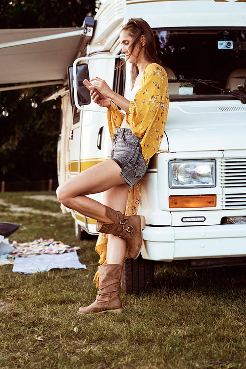 Full length image of young caucasian woman leaning on camper car and browsing mobile phone