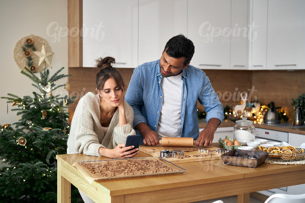 Multi ethnicity couple baking Christmas cookies and looking at mobile phone