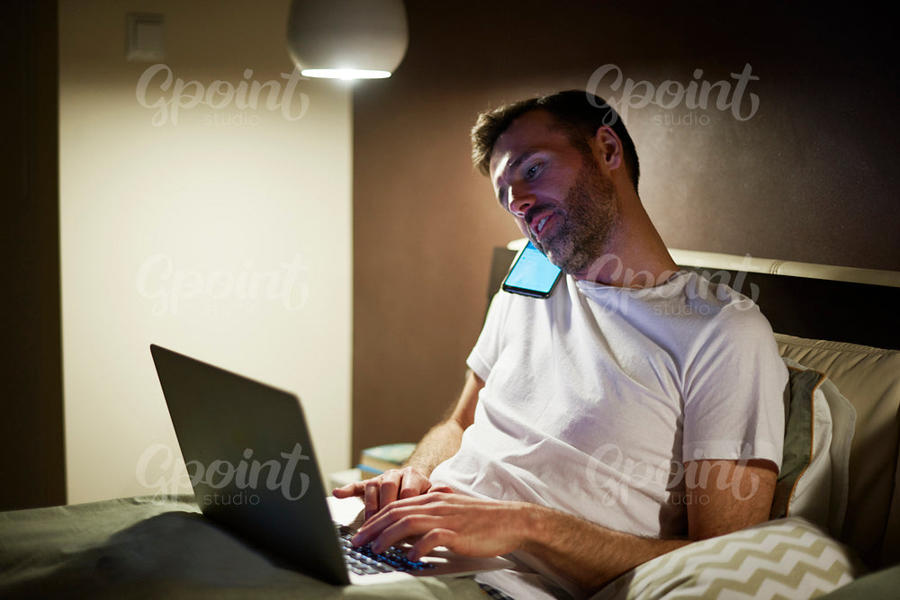 Man talking by mobile phone and using laptop in bed