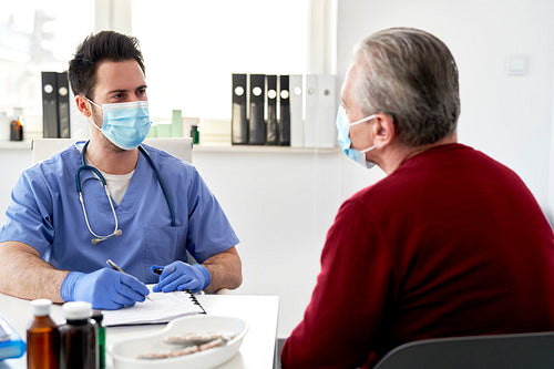 Young doctor in protective mask talking with a senior patient