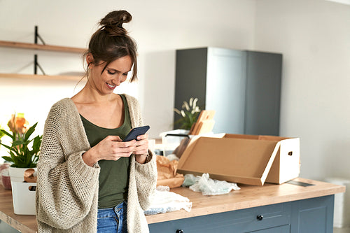 Young caucasian woman using mobile phone while unpacking stuff in new house
