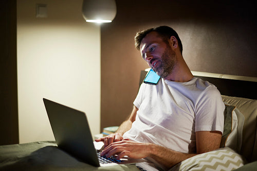 Man talking by mobile phone and using laptop in bed