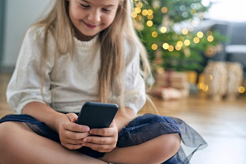 Caucasian girl having fun while browsing smart phone and Christmas tree in the background