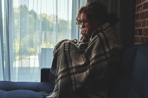 Depressed young caucasian woman sitting on sofa in silence covered with blanket