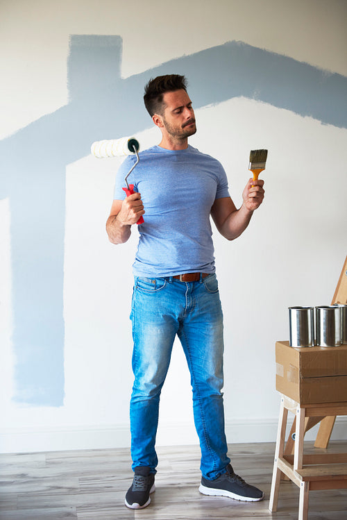 Young man choosing between paint roller and paintbrush