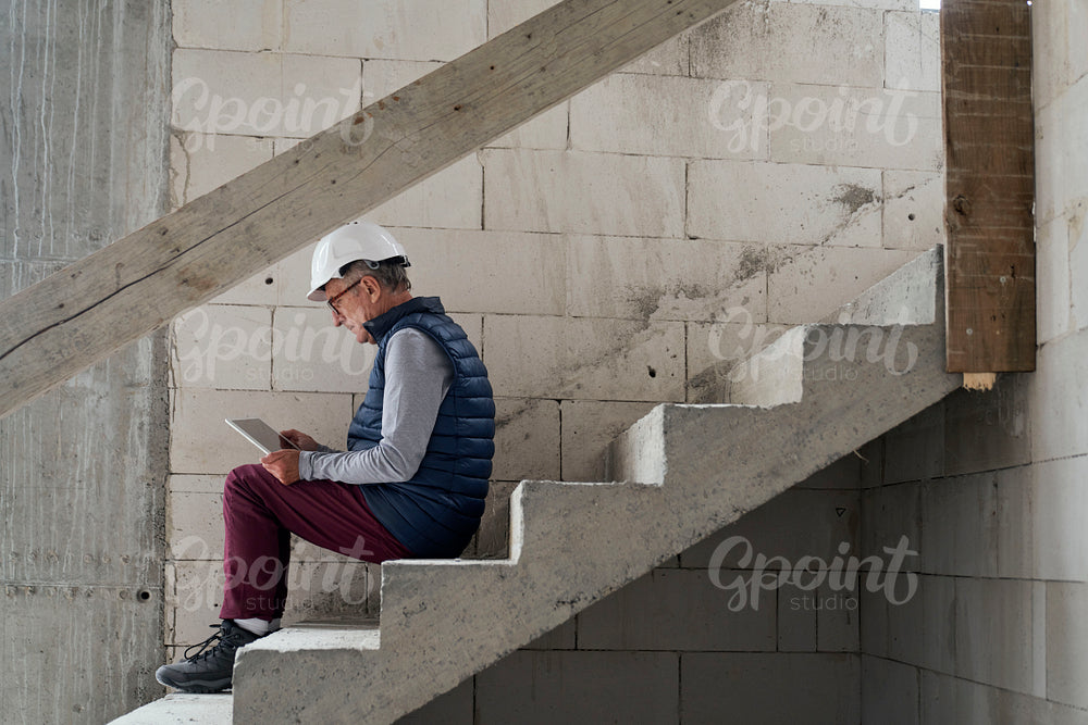 Caucasian engineer sitting on stairs and browsing digital tablet on construction site