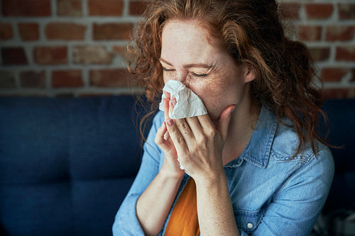 Young caucasian  red head woman blowing nose into tissue at home